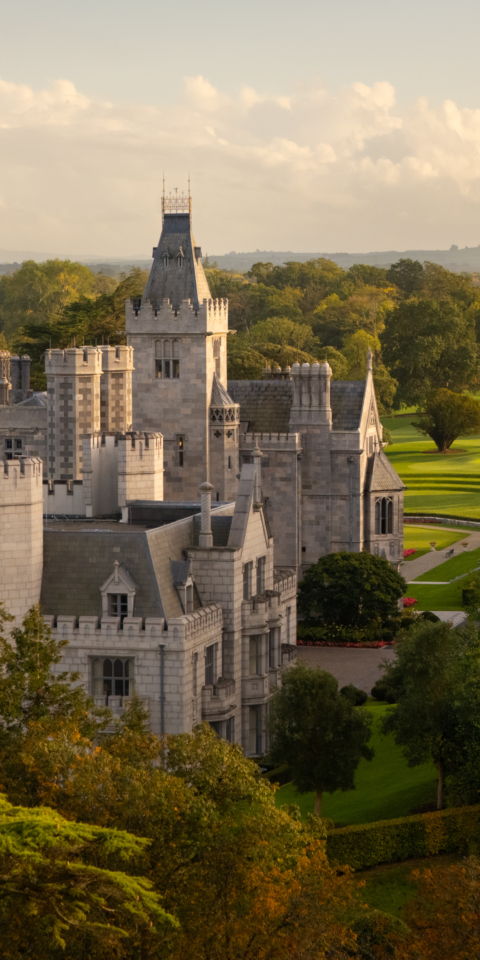 A grand manor house surrounded by lush green lawns and trees, with a majestic architectural design featuring multiple turrets and intricate stonework, set against a cloudy sky.
