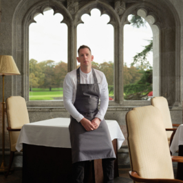 A chef, dressed in white, skillfully plates a gourmet dish in a professional kitchen setting. Sophisticated cooking tools and fresh ingredients are visible in the background.