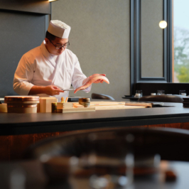 Chef holding a gourmet dish, standing in Oku Restaurant at Adare Manor. The elegant dining setting features wooden furnishings and atmospheric lighting.