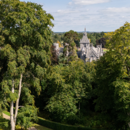 Aerial view of an expansive luxury estate with a grand manor house, manicured gardens, wooded areas, and a golf course under a clear blue sky.