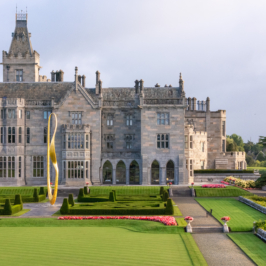 Aerial view of Adare Manor, showing its expansive manicured lawns, intricate architecture, and surrounding dense forests under a clear blue sky.