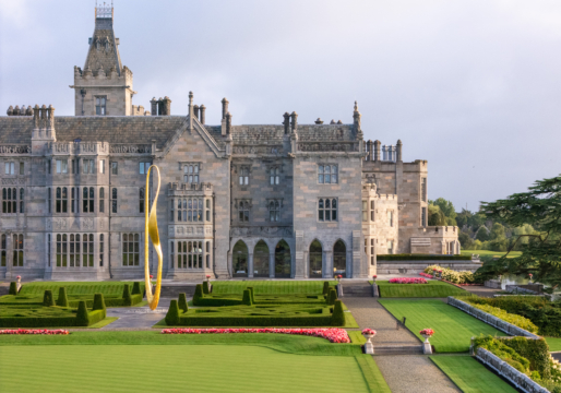 Aerial view of Adare Manor, showing its expansive manicured lawns, intricate architecture, and surrounding dense forests under a clear blue sky.