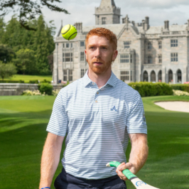 Red‑haired golfer practising with a hurley and sliotar on manicured lawn before Adare Manor’s Gothic Revival façade at Adare, County Limerick, Ireland — 5‑star luxury golf resort, spa and fine dining.