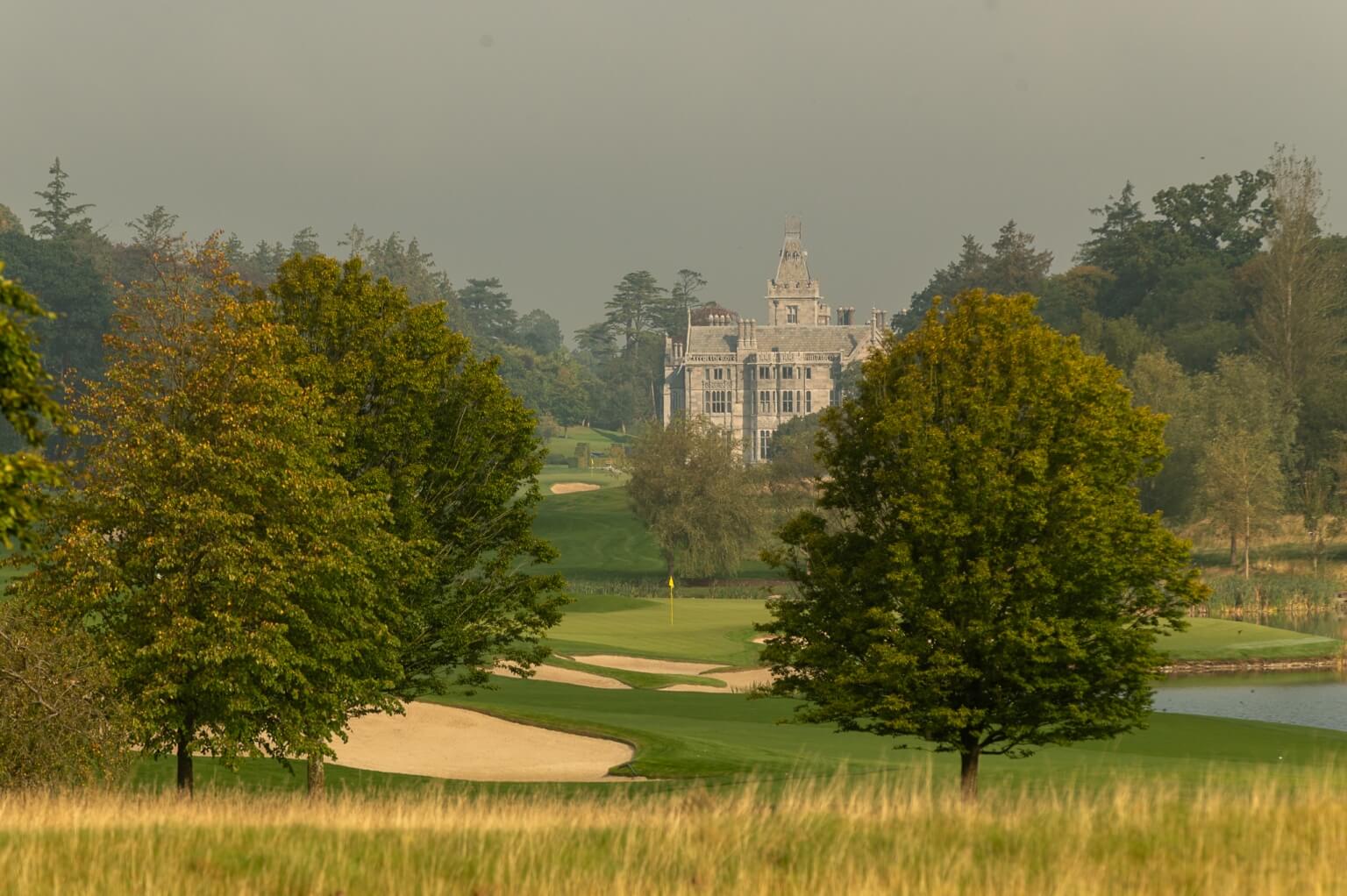 Panoramic view of Adare Manor's Gothic Revival mansion beyond a manicured golf fairway and sand bunkers, golfers on the green amid autumn trees, Adare, County Limerick, Ireland — 5‑star luxury golf resort, spa and fine dining.